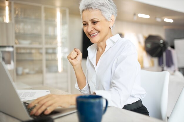 Emocionada empresaria de mediana edad elegante usando la computadora portátil para el trabajo, apretando los puños, feliz de ganar licitación