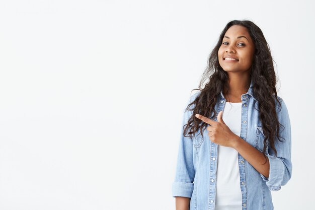 Emocionada y fascinada joven estudiante de piel oscura con camisa azul claro y cabello largo y ondulado que indica algo genial en la pared blanca con espacio para copiar su texto o contenido promocional.