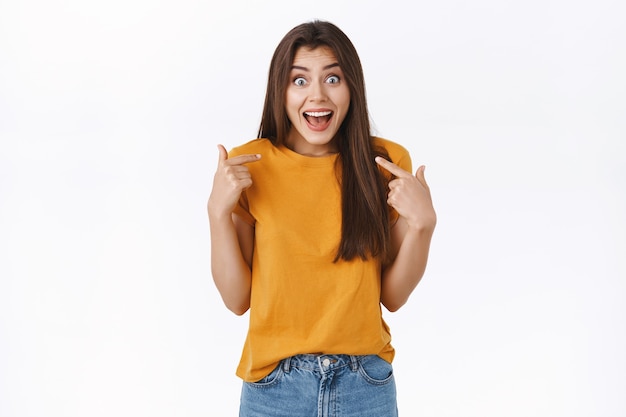 Emocionada, feliz joven ganando algo, mirada sorprendida, no puedo creer su suerte, señalando a sí misma, sonriendo con la boca abierta de asombro de pie sin palabras y emocionada, fondo blanco