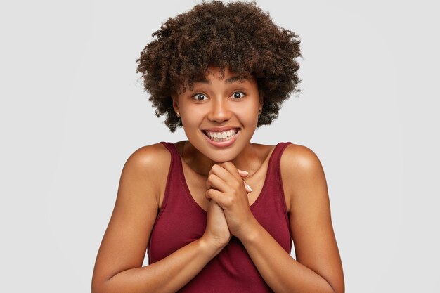 Emocionada feliz joven mujer afro mantiene las manos juntas, reacciona alegremente en un regalo agradable, sonríe ampliamente