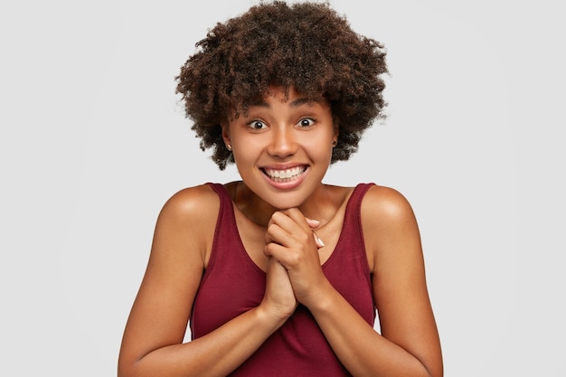 Emocionada feliz joven mujer afro mantiene las manos juntas, reacciona alegremente en un regalo agradable, sonríe ampliamente