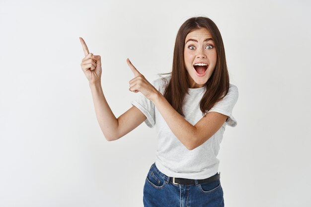 Emocionada, feliz y sorprendida hermosa joven, preste atención a la pancarta, señalando con el dedo hacia la izquierda y gritando de alegría, párese en la pared blanca