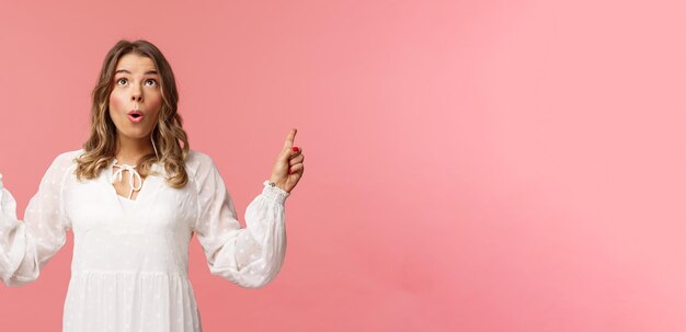 Emocionada hermosa mujer rubia caucásica con vestido blanco doblar los labios con emoción mirando apuntando con el dedo