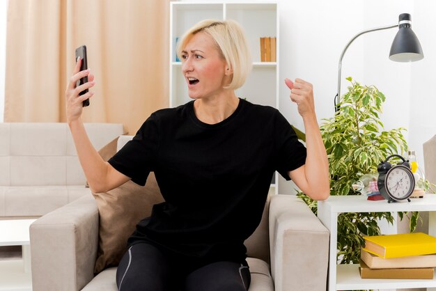 Emocionada hermosa mujer rusa rubia sentada en un sillón manteniendo el puño y mirando el teléfono