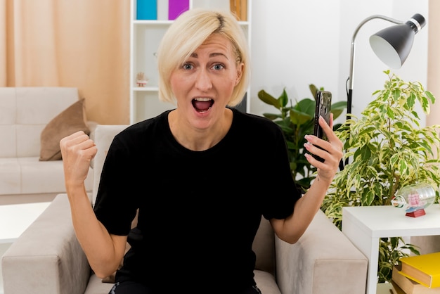 Emocionada hermosa mujer rusa rubia sentada en un sillón manteniendo el puño y sosteniendo el teléfono mirando a la cámara dentro de la sala de estar