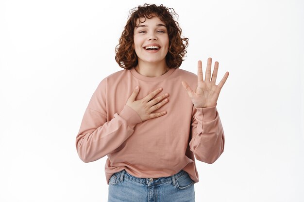 Emocionada hermosa niña sonriendo, pidiendo participar diciendo hola, mencionándose a sí misma, levantando la mano y colocando la palma sobre el corazón, siendo honesta, de pie en blanco.