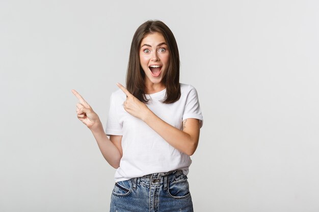 Emocionada hermosa niña sonriendo, señalando con el dedo la esquina superior izquierda y mirando complacida al logo.