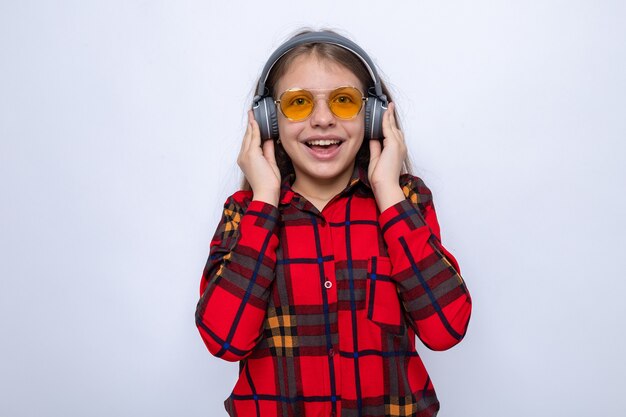 Emocionada hermosa niña vestida con camisa roja y gafas con auriculares aislados en la pared blanca