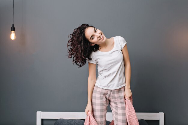 Emocionada joven alegre con cabello rizado morena en pijama divirtiéndose en la cama. Sonriendo, expresando verdaderas emociones positivas, relajándose en casa en un apartamento moderno