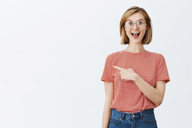 Emocionada joven atractiva con gafas mostrando el camino, apuntando con el dedo a la izquierda en copyspace
