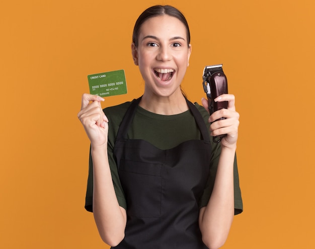 Emocionada joven barbero morena en uniforme sostiene tiene cortapelos y tarjeta de crédito en naranja
