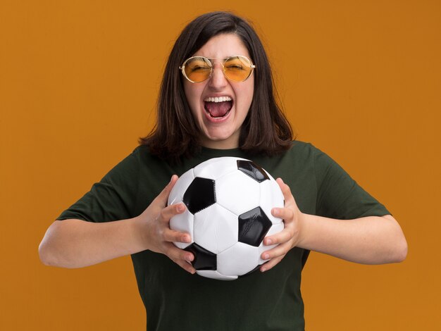 Emocionada joven bastante caucásica en gafas de sol sosteniendo la bola aislada en la pared naranja con espacio de copia