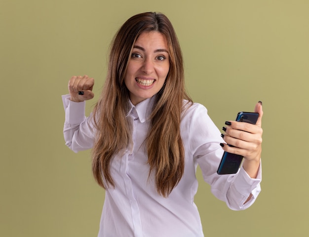 Emocionada joven bastante caucásica mantiene el puño y sostiene el teléfono aislado en la pared verde oliva con espacio de copia