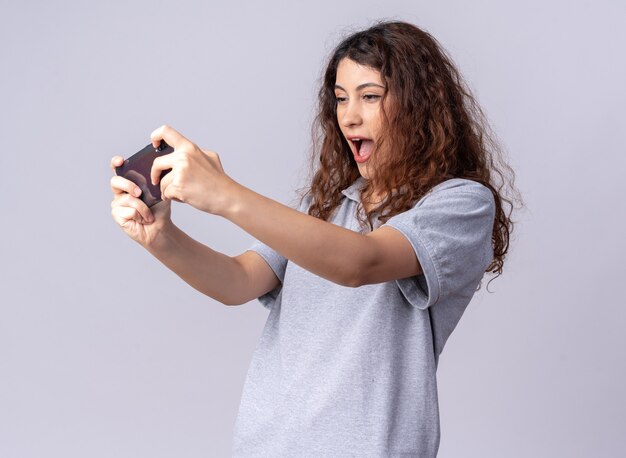 Emocionada joven bastante caucásica de pie en la vista de perfil sosteniendo el teléfono móvil jugando en el teléfono aislado en la pared blanca