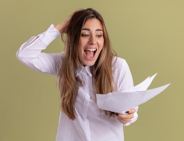 Foto gratuita emocionada joven bastante caucásica pone la mano en la cabeza sosteniendo y mirando hojas de papel en blanco en verde oliva