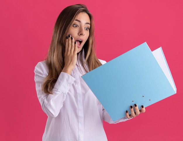 Emocionada joven bastante caucásica pone la mano en la cara sosteniendo y mirando la carpeta de archivos