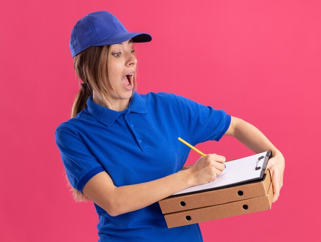 Emocionada joven bonita repartidora en uniforme sostiene cajas de pizza y escribe en el portapapeles con bolígrafo en rosa