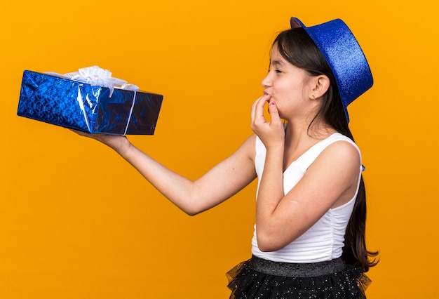 Emocionada joven caucásica con gorro de fiesta azul sosteniendo y mirando la caja de regalo poniendo la mano en la boca aislada en la pared naranja con espacio de copia