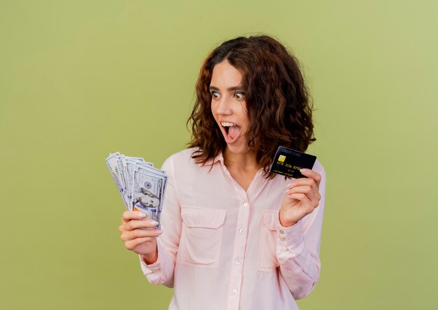 Foto gratuita emocionada joven caucásica tiene tarjeta de crédito y mira dinero aislado sobre fondo verde con espacio de copia