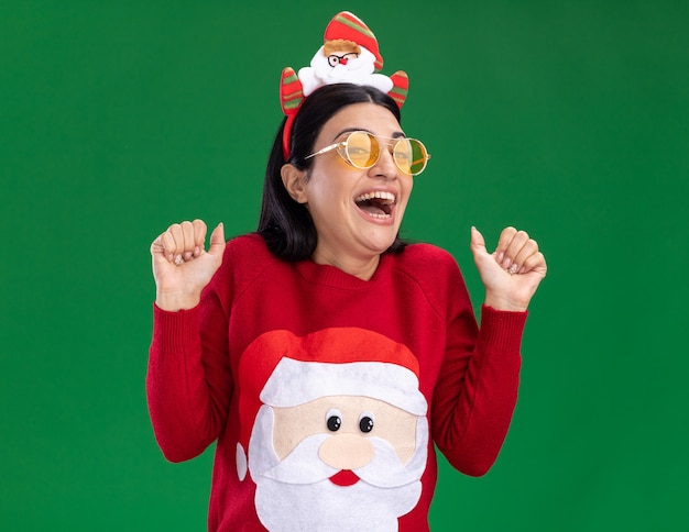 Foto gratuita emocionada joven caucásica vistiendo suéter y diadema de santa claus con gafas mirando a la cámara mostrando los pulgares para arriba aislado sobre fondo verde