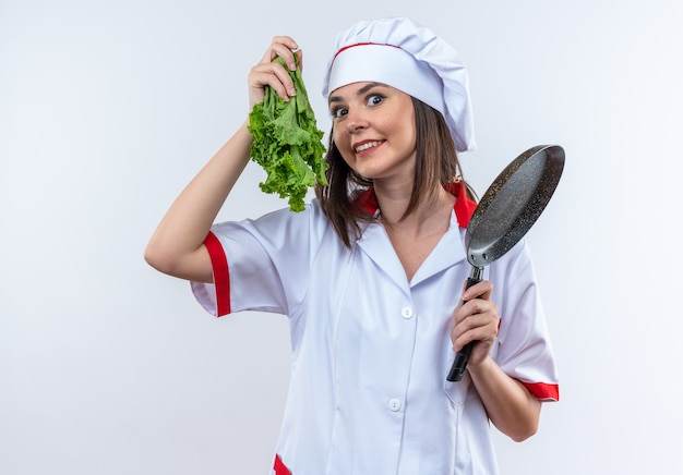 Emocionada joven cocinera vistiendo uniforme de chef sosteniendo ensalada con sartén aislado sobre fondo blanco.