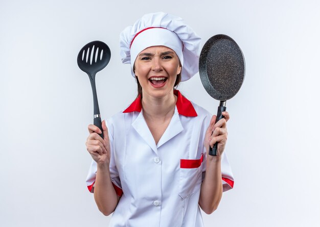 Emocionada joven cocinera vistiendo uniforme de chef sosteniendo una espátula con sartén aislado sobre fondo blanco.