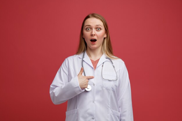 Emocionada joven doctora rubia vistiendo bata médica y un estetoscopio alrededor del cuello apuntando a sí misma