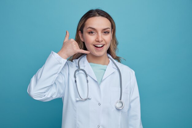 Emocionada joven doctora vistiendo bata médica y estetoscopio alrededor del cuello haciendo gesto de llamada