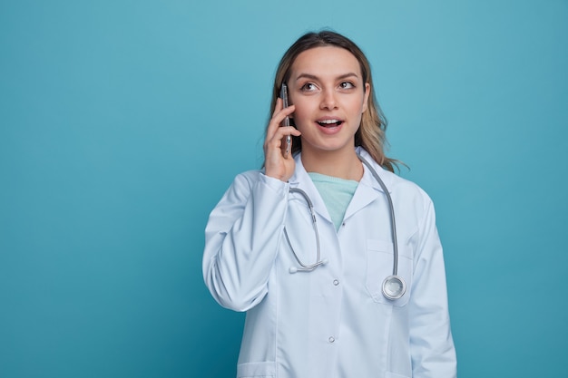 Emocionada joven doctora vistiendo bata médica y un estetoscopio alrededor del cuello mirando hacia arriba hablando por teléfono