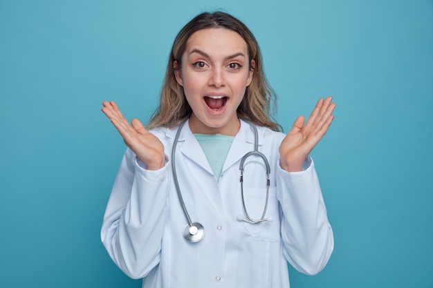 Emocionada joven doctora vistiendo bata médica y estetoscopio alrededor del cuello mostrando las manos vacías