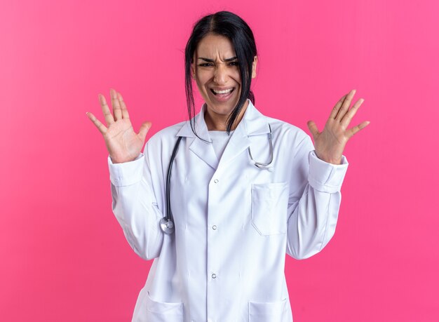 Emocionada joven doctora vistiendo bata médica con estetoscopio extendiendo las manos aisladas en la pared rosa