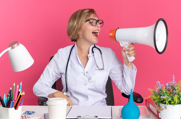 Emocionada joven doctora vistiendo bata médica con estetoscopio y gafas se sienta en el escritorio con herramientas médicas habla por altavoz aislado en la pared rosa