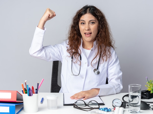 Emocionada joven doctora vistiendo bata médica y un estetoscopio sentado en la mesa con herramientas médicas manteniendo la mano sobre la mesa mirando al frente haciendo un gesto fuerte aislado en la pared blanca