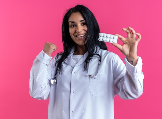 Emocionada joven doctora vistiendo bata médica con estetoscopio sosteniendo pastillas mostrando gesto sí aislado en la pared rosa