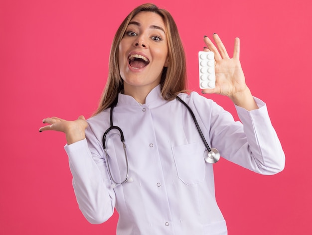 Emocionada joven doctora vistiendo bata médica con estetoscopio sosteniendo píldoras puntos detrás aislado en pared rosa