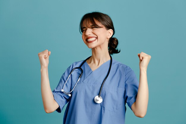 Emocionada joven doctora vistiendo uniforme fith estetoscopio aislado sobre fondo azul.