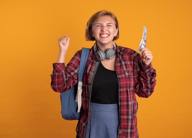 Emocionada joven estudiante eslava con auriculares con mochila se encuentra con los ojos cerrados levantando el puño sosteniendo dinero