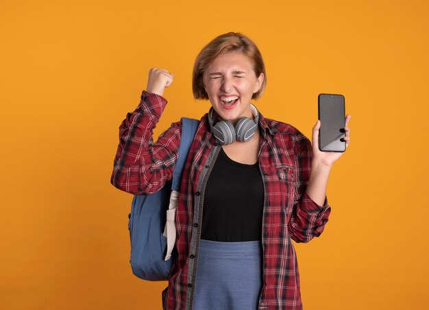 Emocionada joven estudiante eslava con auriculares con mochila se encuentra con los ojos cerrados levantando el puño sosteniendo el teléfono