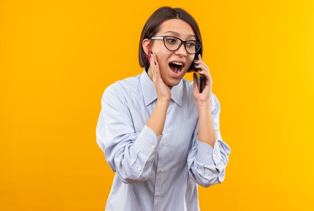 Emocionada joven hermosa con gafas habla por teléfono poniendo la mano en la mejilla aislada en la pared naranja