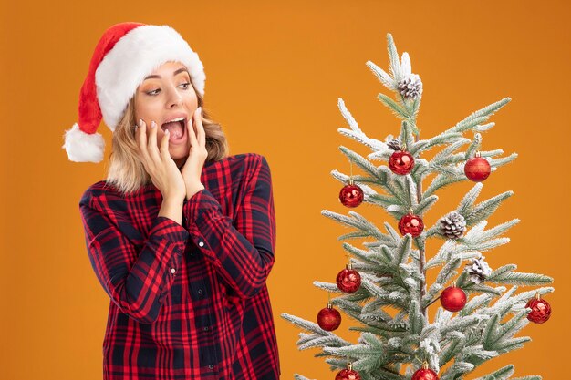 Emocionada joven hermosa de pie cerca del árbol de Navidad con sombrero de Navidad poniendo las manos en las mejillas aisladas sobre fondo naranja