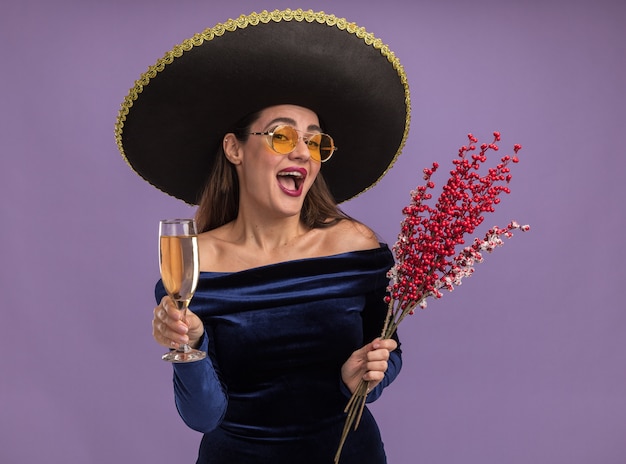 Emocionada joven hermosa vestida con vestido azul y gafas con sombrero sosteniendo la rama de serbal con copa de champagneisolated sobre fondo púrpura