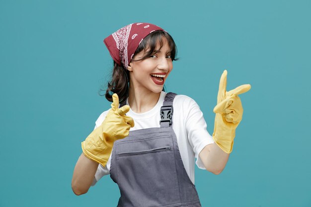 Emocionada joven limpiadora con pañuelo uniforme y guantes de goma mirando a la cámara mostrándote un gesto aislado en el fondo azul