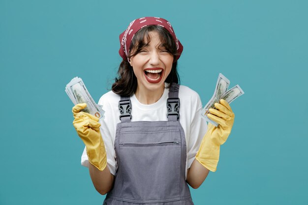 Emocionada joven limpiadora con pañuelo uniforme y guantes de goma sosteniendo dinero en ambas manos gritando mientras miraba la cámara aislada en el fondo azul
