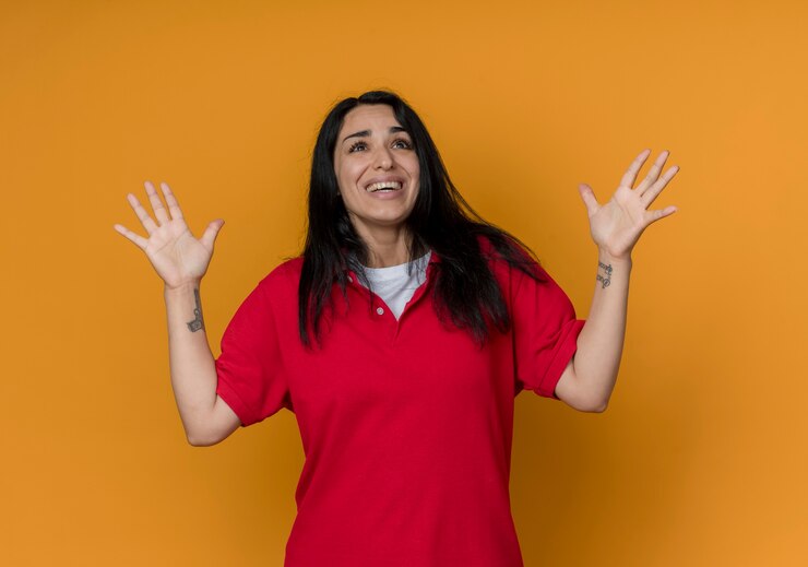 Foto gratuito emocionada joven morena caucásica vestida con camisa roja levanta las manos mirando hacia arriba aislado en la pared naranja