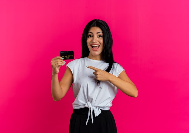 Emocionada joven mujer caucásica sostiene y apunta a la tarjeta de crédito mirando a la cámara