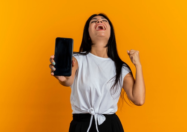 Emocionada joven mujer caucásica sostiene el teléfono y mantiene el puño mirando hacia arriba