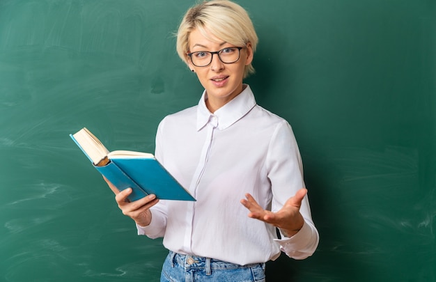 Emocionada joven profesora rubia con gafas en el aula de pie frente a la pizarra sosteniendo un libro que muestra la mano vacía con espacio de copia