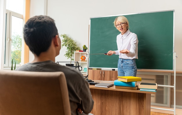 Foto gratuita emocionada joven profesora rubia con gafas en el aula de pie frente a la pizarra sosteniendo un puntero mirando sentado estudiante adolescente chico mostrando la mano vacía