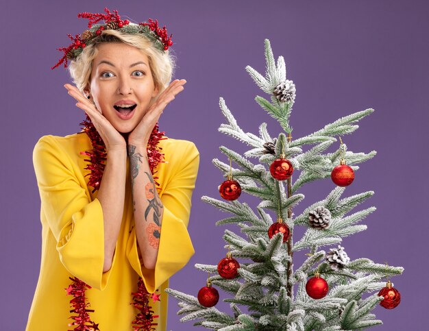 Emocionada joven rubia con corona de Navidad y guirnalda de oropel alrededor del cuello de pie cerca del árbol de Navidad decorado manteniendo las manos en la cara mirando a cámara aislada sobre fondo púrpura