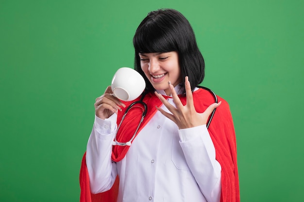 Emocionada joven superhéroe con estetoscopio con bata médica y manto sosteniendo y mirando la taza aislada en verde